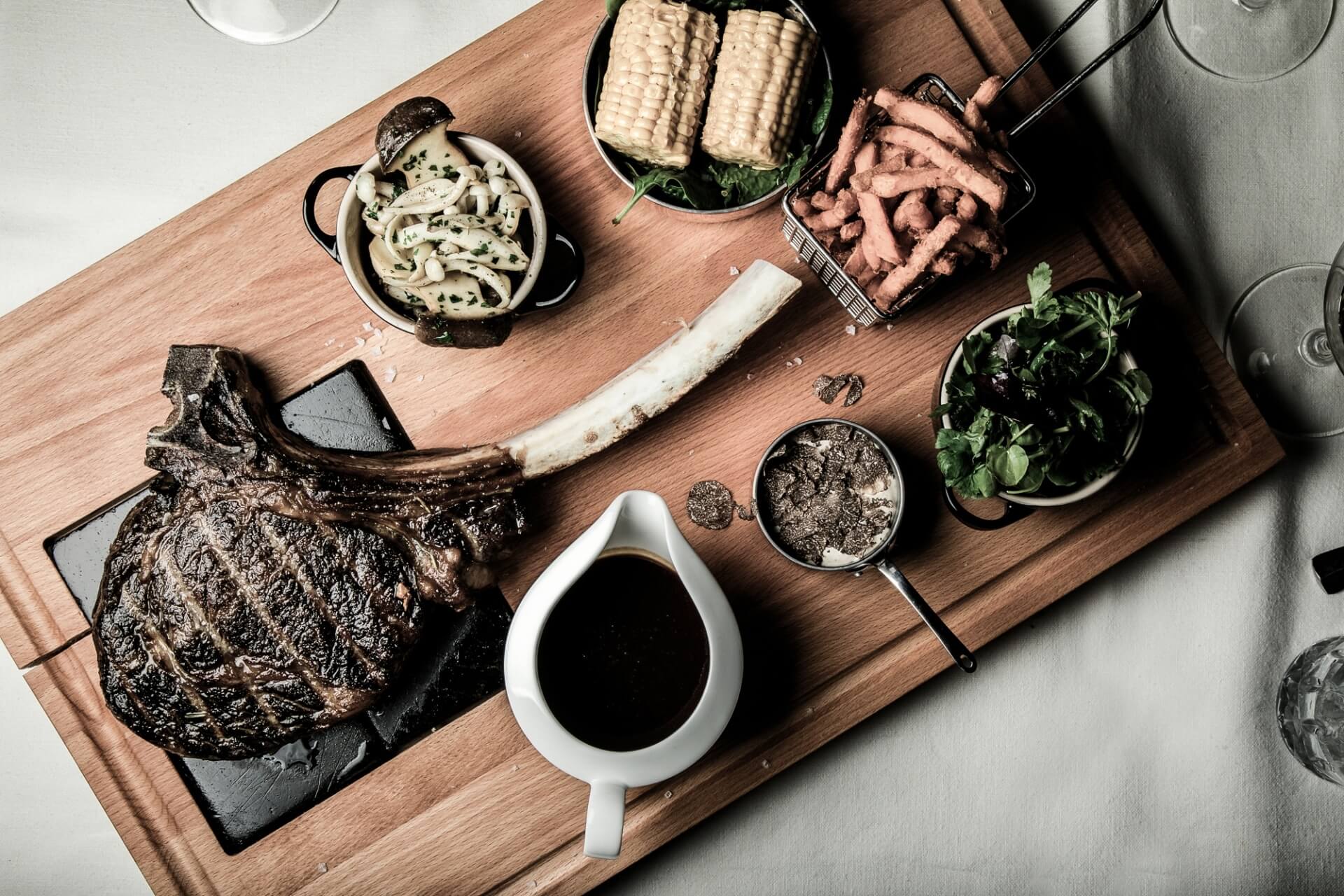 Ein üppiges Festmahl mit einem großen gegrillten Tomahawk-Steak, dazu Maiskolben, Zwiebelringe, Salat und Sahnesauce, elegant präsentiert auf einem Holzbrett in gehobener Atmosphäre.