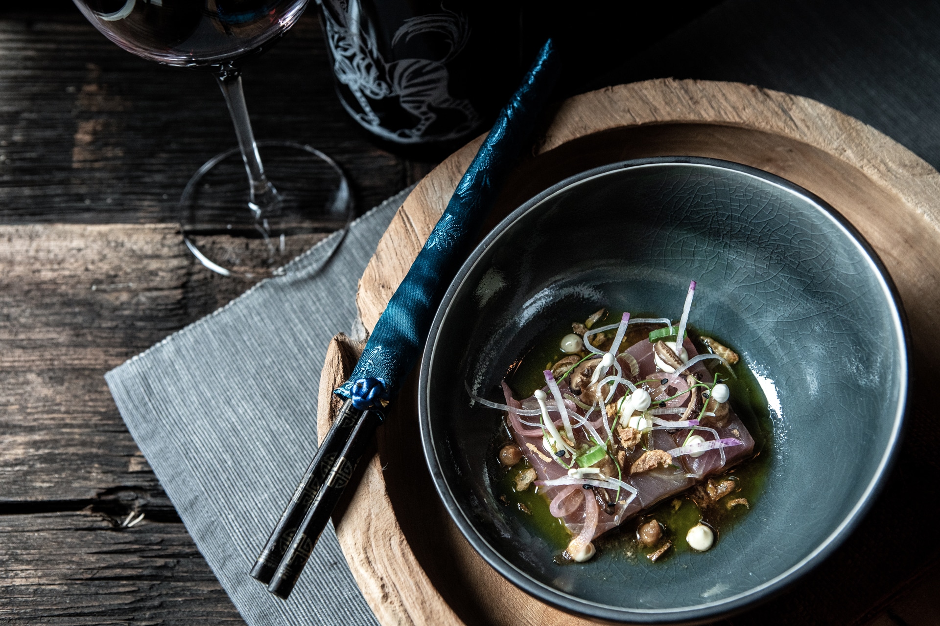 Elegant fine dining presentation with an artistic dish of delicately arranged sashimi served in a dark bowl, complemented by a pair of blue chopsticks on a wooden rest, all set on a rustic wooden table beside a glass of red wine.