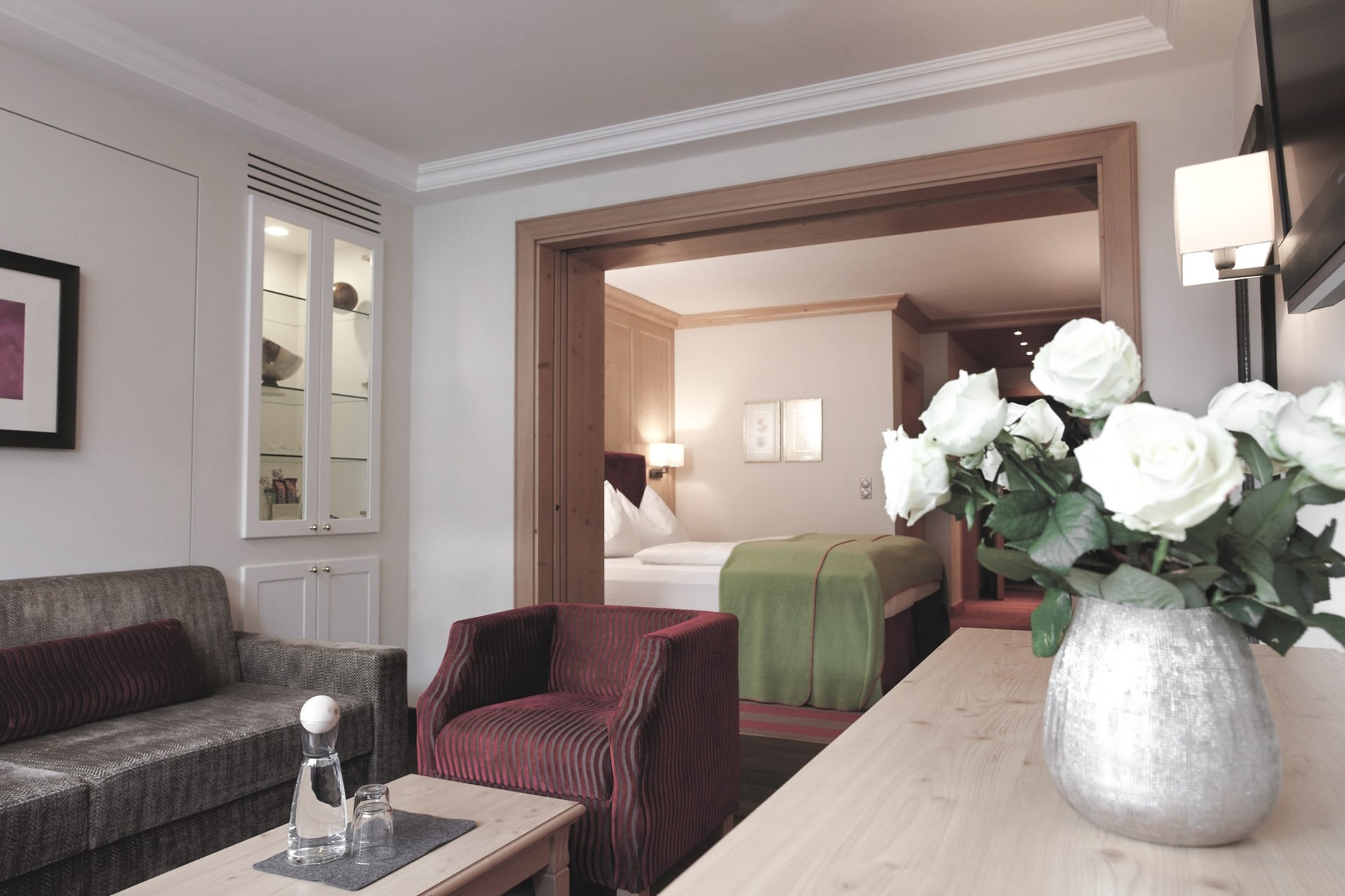 Elegantes und gemütliches Interieur einer Hotelsuite mit einem geschmackvoll eingerichteten Wohnbereich, der in ein bequemes Schlafzimmer übergeht, akzentuiert mit frischen weißen Rosen auf einem Holztisch für einen Hauch von Klasse.