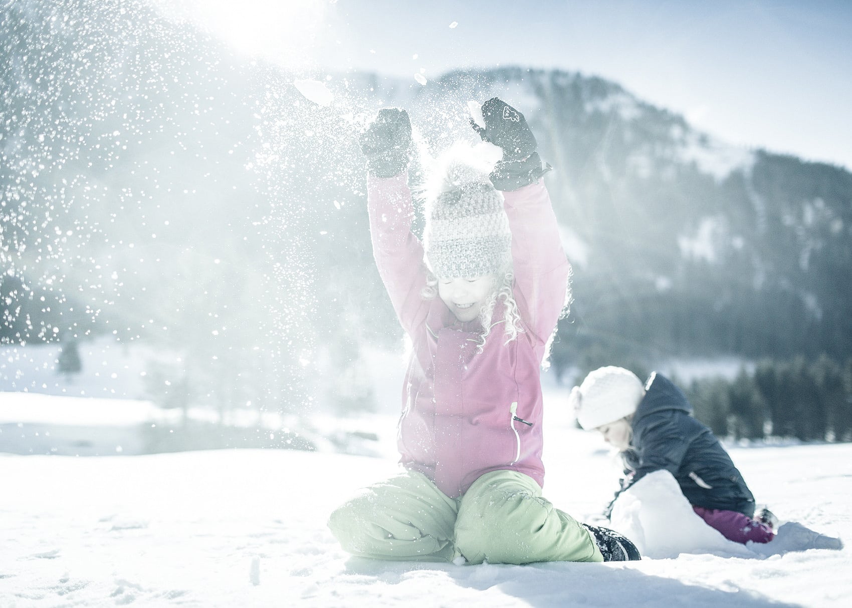 A joyful child playing with snow, tossing it up into the air on a sunny winter day while another child is seen building something with snow in the background.