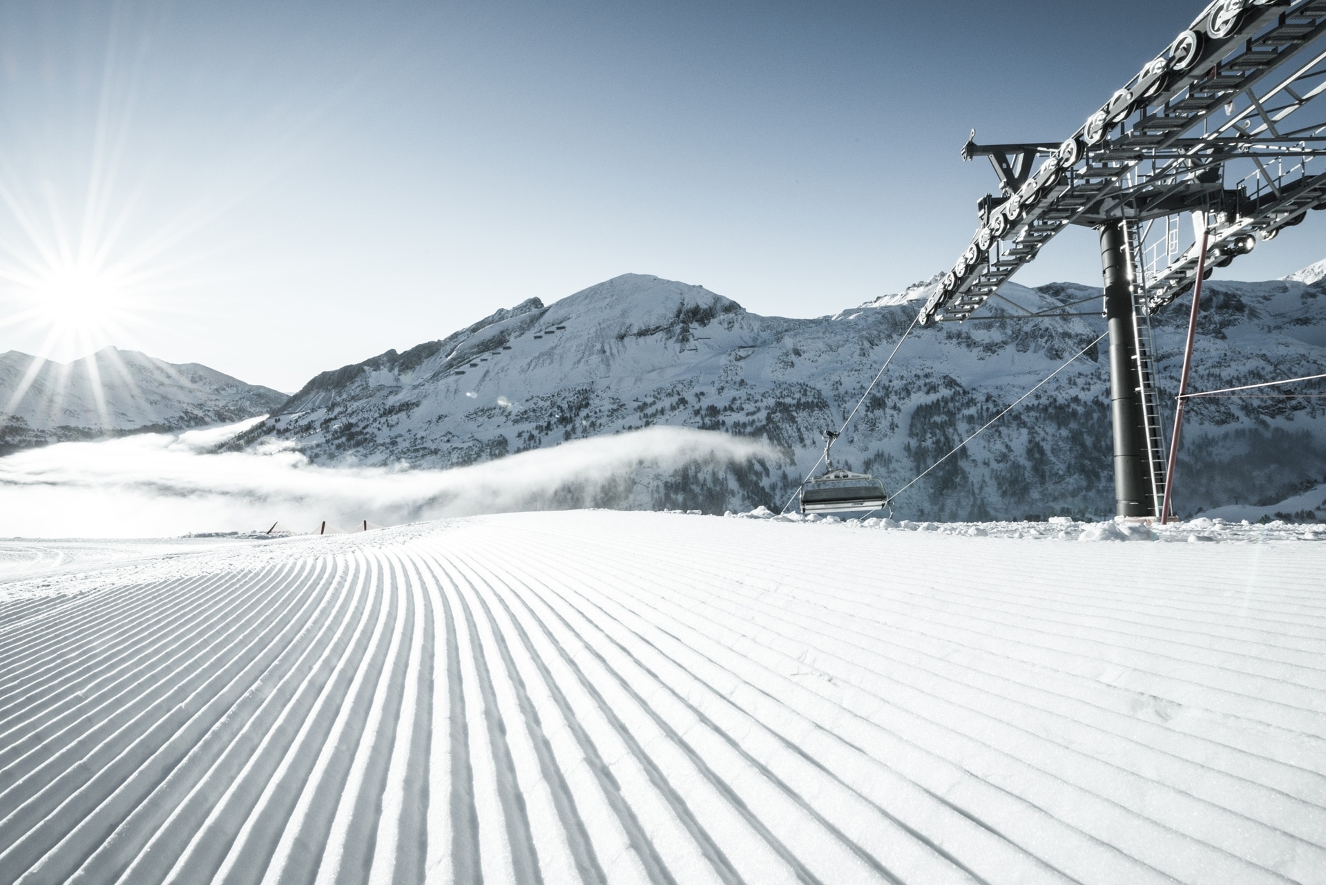 Sunrise over a pristine ski slope with freshly groomed tracks and a ski lift, set against a backdrop of majestic snow-covered mountains.