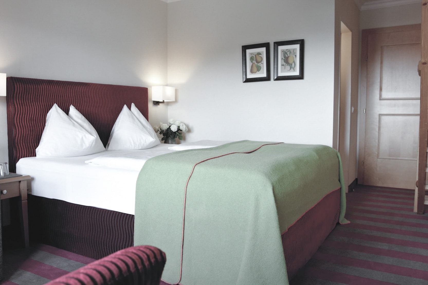 Ein ordentlich eingerichtetes Hotelzimmer mit einem großen Bett, das mit einer grünen Tagesdecke, frischer weißer Bettwäsche und weichen Kissen ausgestattet ist, ergänzt durch einen gestreiften Teppich und gerahmte Kunst an der Wand, die ein Gefühl von Komfort und Einfachheit vermitteln.