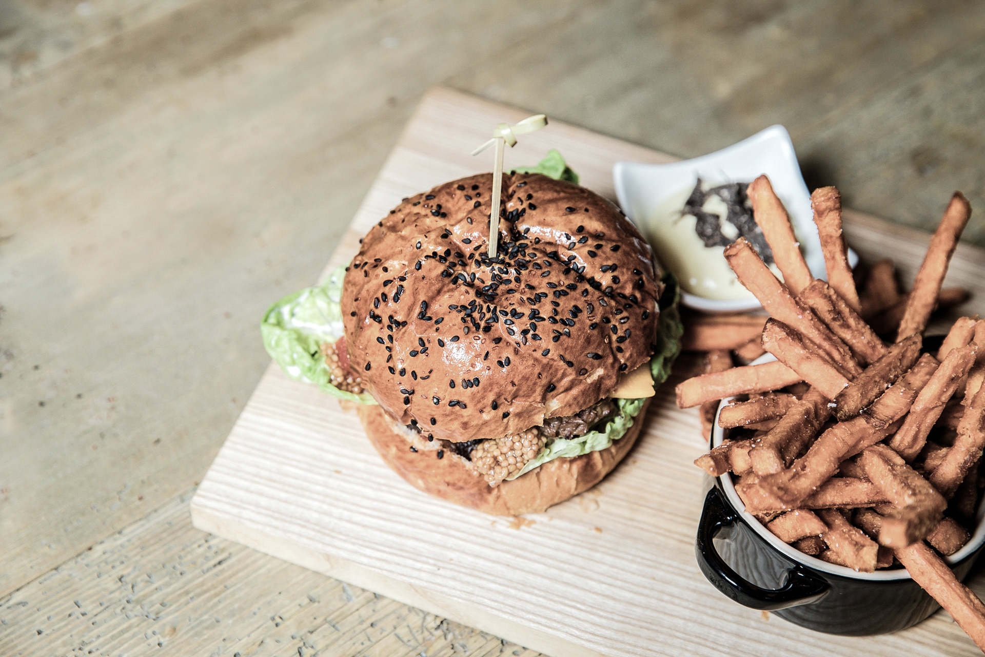 A delicious gourmet burger with sesame seed bun and fresh lettuce, accompanied by a side of crispy sweet potato fries, served on a rustic wooden cutting board.