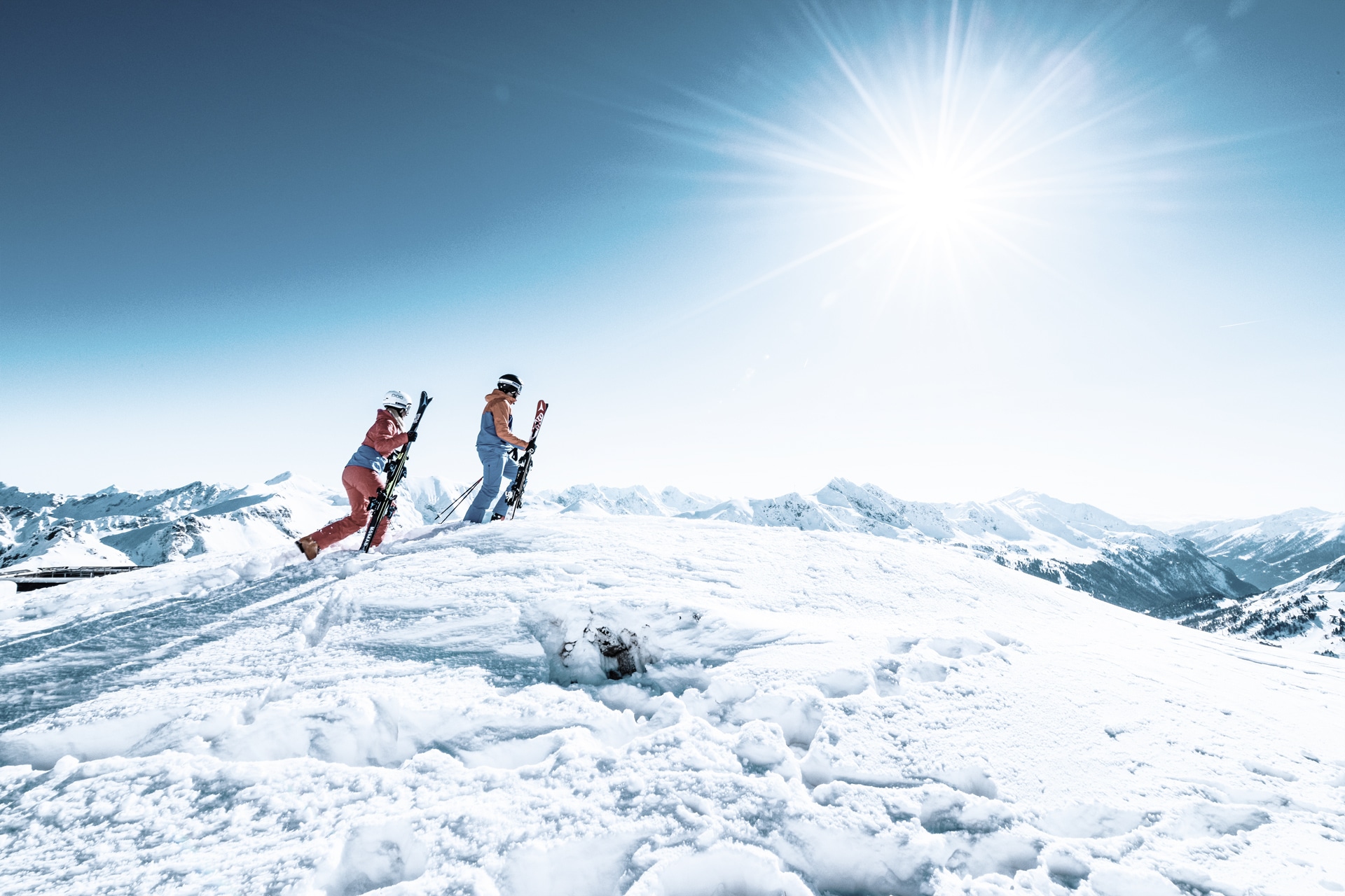 Two skiers on a snowy mountain peak with clear blue skies, basking in the radiant sunlight while preparing for an exhilarating descent.