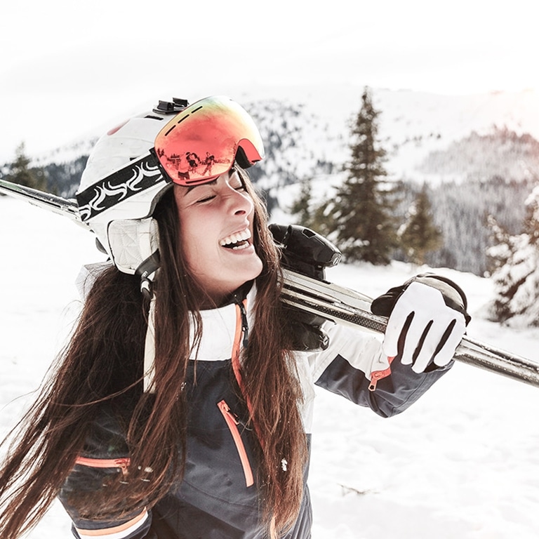 Eine fröhliche Skifahrerin mit ihren Skiern auf der Schulter genießt einen sonnigen Wintertag auf der Piste vor der Kulisse schneebedeckter Berge.