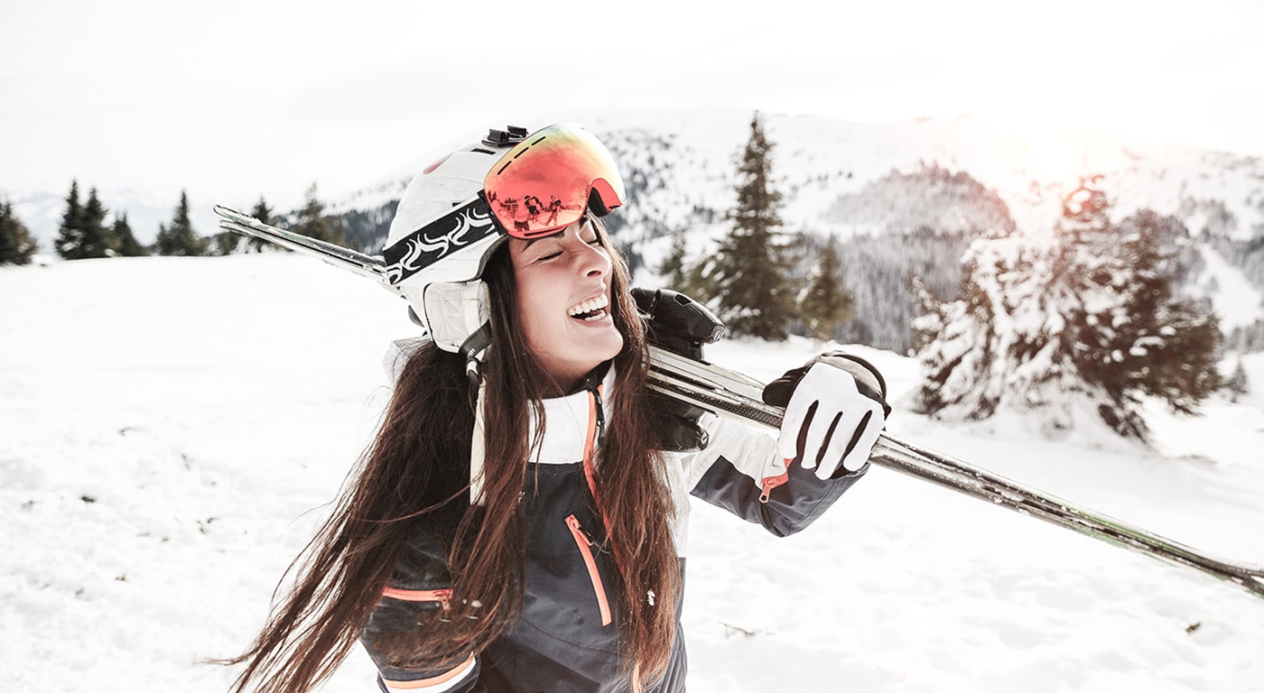 Eine fröhliche Skifahrerin mit ihren Skiern auf der Schulter genießt einen sonnigen Wintertag auf der Piste vor der Kulisse schneebedeckter Berge.