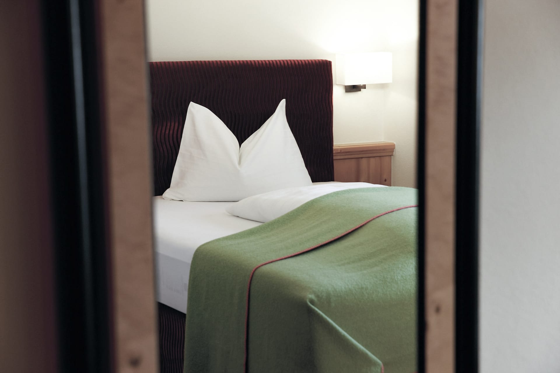 A neatly made bed with white linens and triangular folded pillows reflected in a mirror in a cozy hotel room.