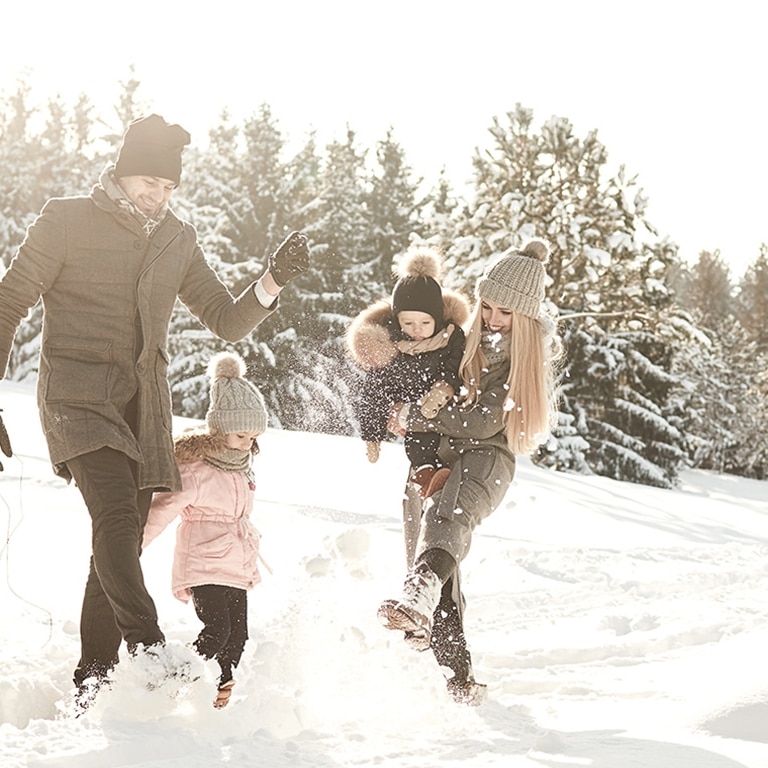 Eine fröhliche Familie verbringt einen lustigen Wintertag: Die Eltern schaukeln ihr Kind spielerisch an den Armen durch eine verschneite Landschaft, während ein anderes Kind aufgeregt zusieht.