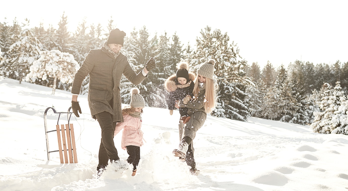 Eine fröhliche Familie verbringt einen lustigen Wintertag: Die Eltern schaukeln ihr Kind spielerisch an den Armen durch eine verschneite Landschaft, während ein anderes Kind aufgeregt zusieht.