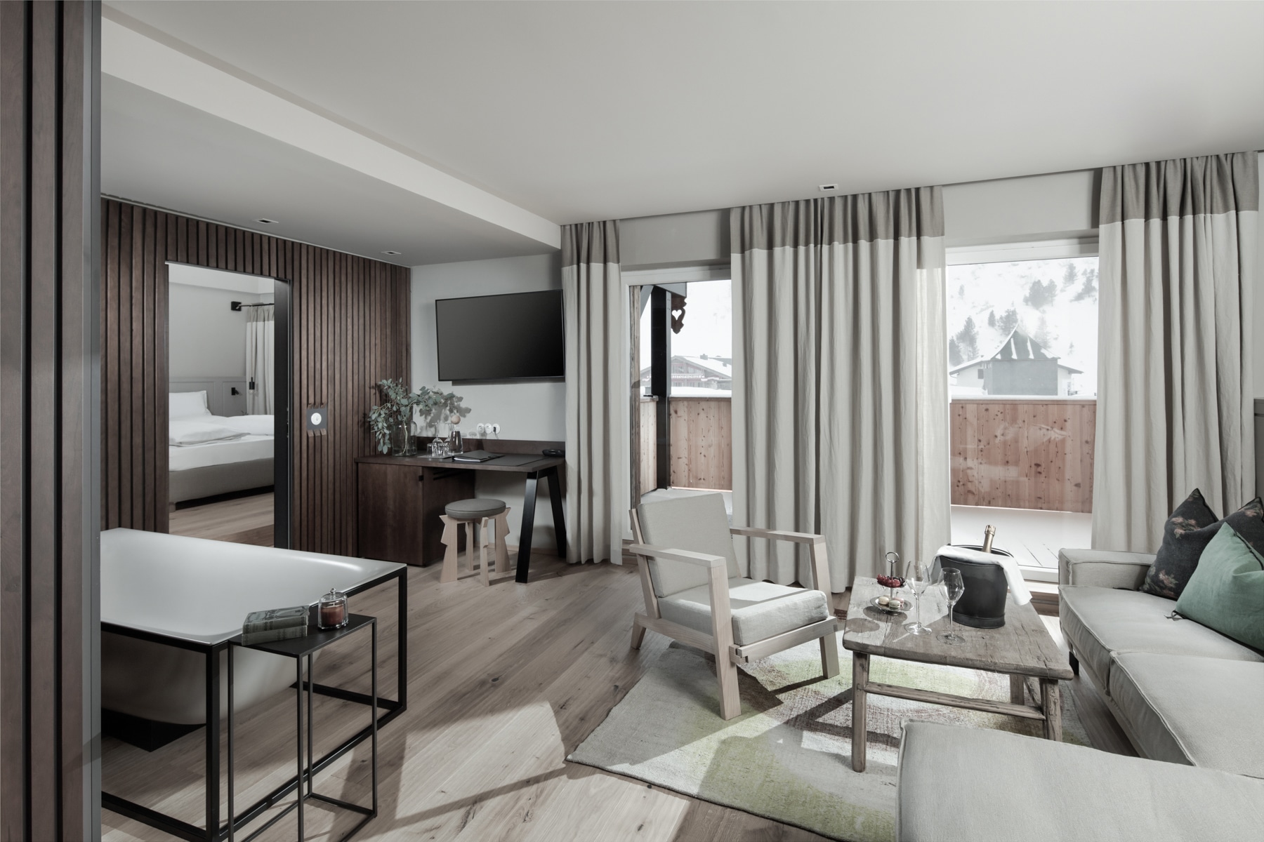 Modern and elegant hotel suite interior with a seamless transition from the inviting living area to the cozy bedroom, featuring neutral tones and contemporary furnishings, offering guests a tranquil retreat with a view of the mountainous landscape.