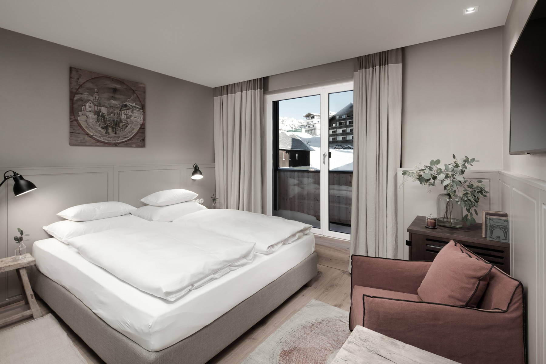 A modern and cozy bedroom featuring a large bed with white linens, an armchair by the window, and tasteful decorations, with a view of urban surroundings.