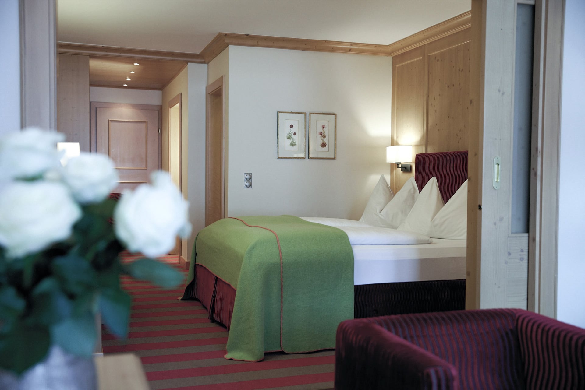 A tranquil and neatly arranged hotel room with warm wooden finishes, soft lighting, and a welcoming bed adorned with a green bedspread and red accents, portraying an inviting atmosphere for rest and relaxation.