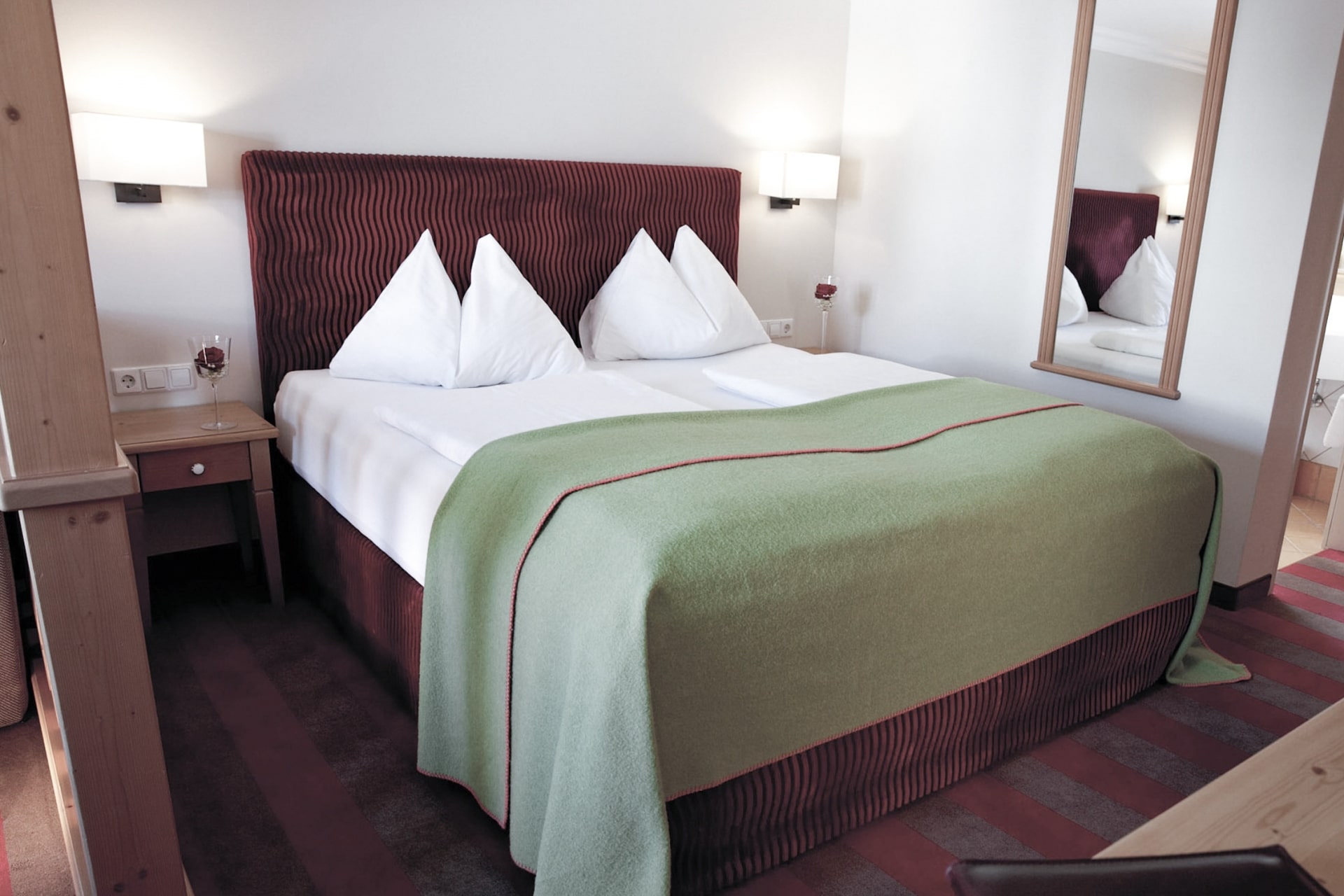 A neatly arranged hotel room with a plush double bed, featuring a burgundy headboard, crisp white sheets, a green bedspread with a red trim, and fluffed white pillows. a bedside table, lamp, and a mirror reflecting part of the room are also visible, creating a cozy and inviting atmosphere.