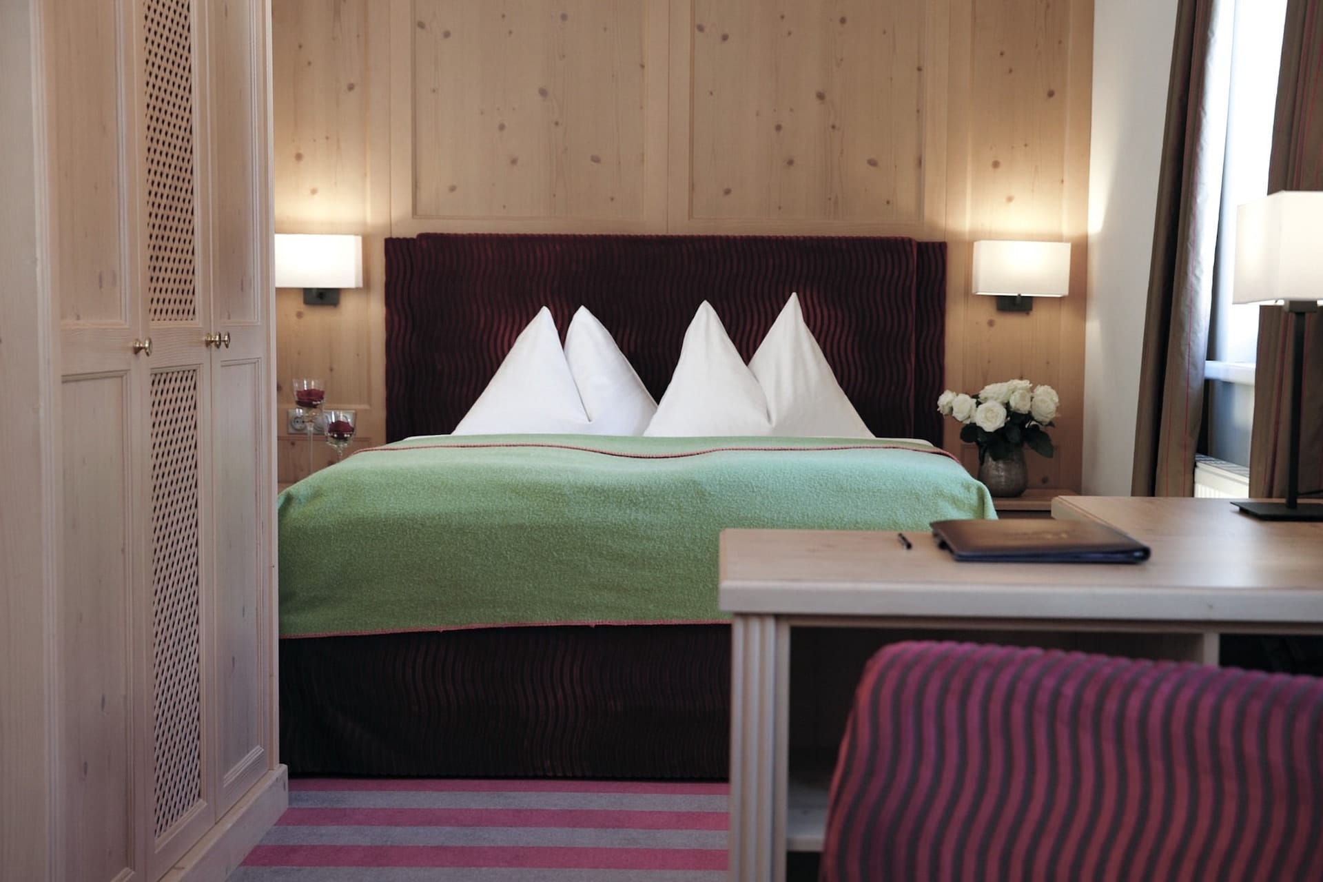 A cozy hotel room featuring a large bed with a green blanket and white, crisply folded pillows, flanked by wooden nightstands and warm lighting, with a work desk situated towards the front. the room's interior design combines modern amenities with a touch of alpine charm embodied by the wooden wall paneling.